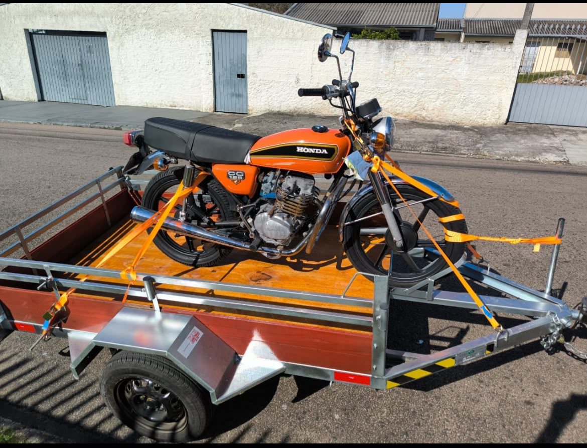 Carretinha de piso de madeira e estrutura galvanizada levando uma moto laranja presa com cintas